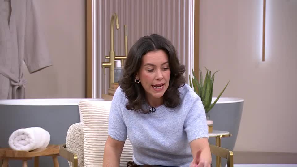 A woman with dark, curly hair is speaking animatedly, gesturing with her right hand. She's seated in a chair, wearing a pale blue sweater, and appears to be on a QVC Beauty segment, likely in the UK.