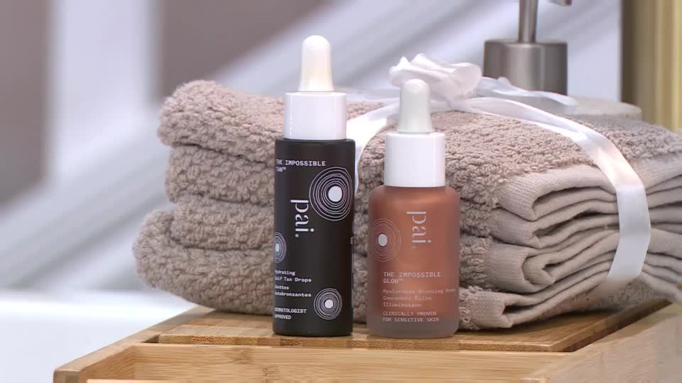 Two Pai skincare bottles sit on a bamboo tray, one dark and the other a warm bronze, beside a stack of neatly folded tan towels. A white ribbon wraps around the towels, and a silver dispenser rises behind them.
