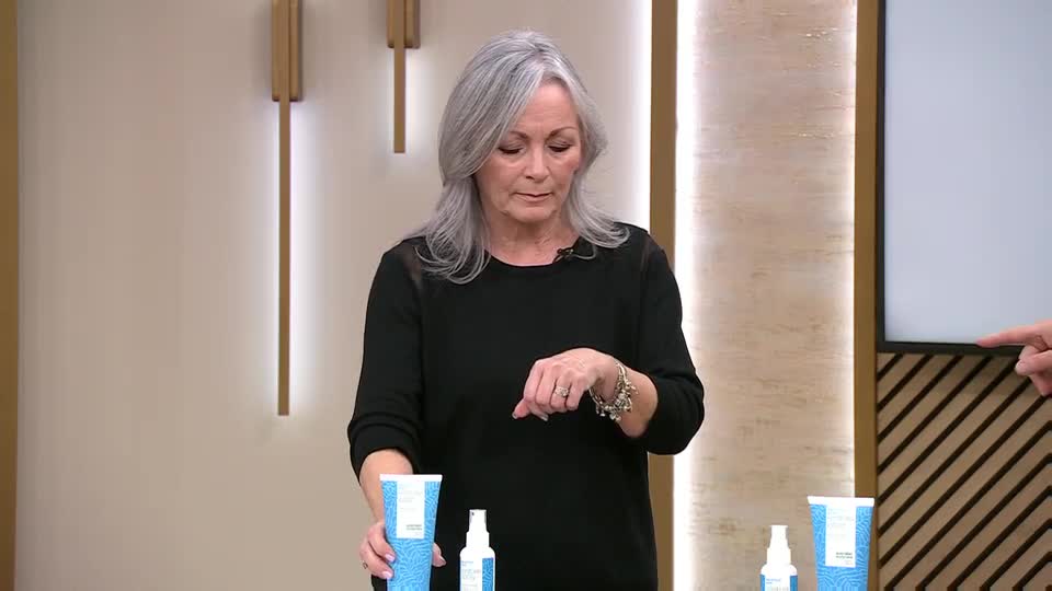 A woman with long grey hair, dressed in black, holds a blue and white tube. She gestures with her right hand, adorned with a bracelet, towards the items on the table.