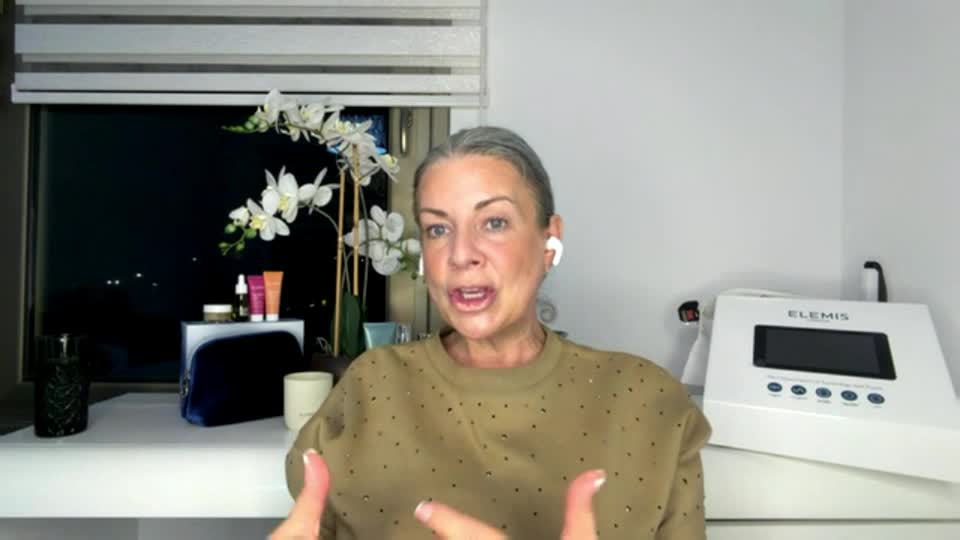 A woman with grey hair, wearing a taupe top, talks animatedly with her hands. Beside her, a white Elemis machine sits on a counter.