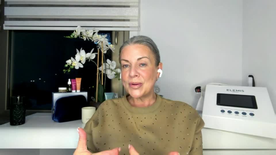 A woman with grey hair, wearing a tan sweater, speaks animatedly. Beside her, a white Elemis device sits on a shelf.