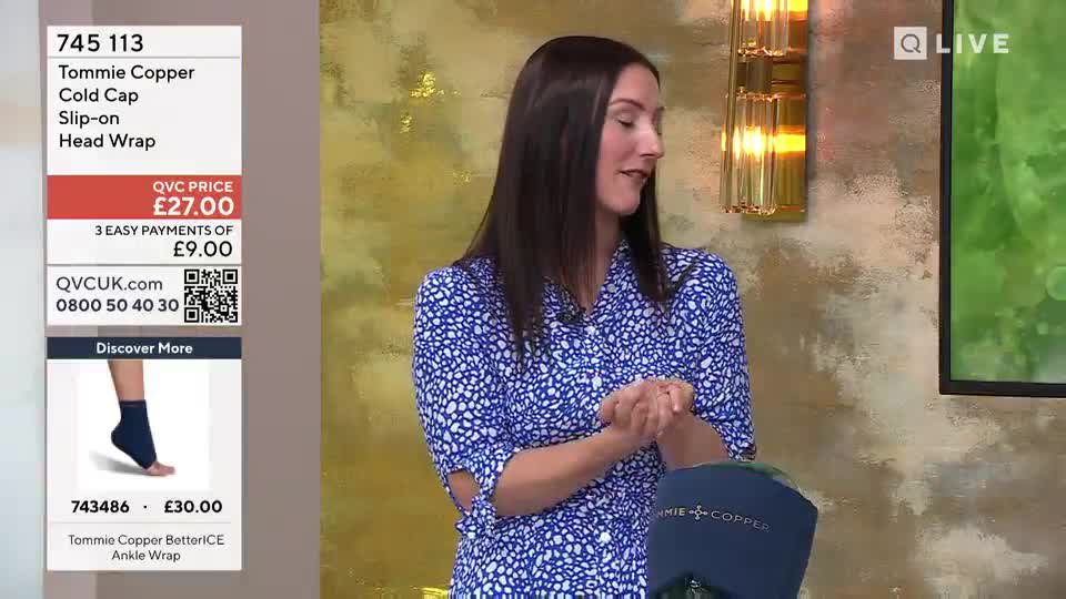 A woman in a blue patterned dress stands on a QVC Beauty set. She gestures with her hands as she talks about a Tommie Copper cold cap.