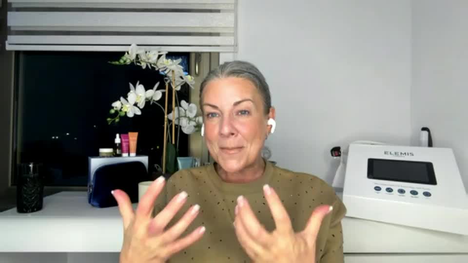 A woman with grey hair, wearing a brown top, gestures with her hands while speaking. Behind her, a white Elemis device sits on a shelf next to a window showing a dark night sky.