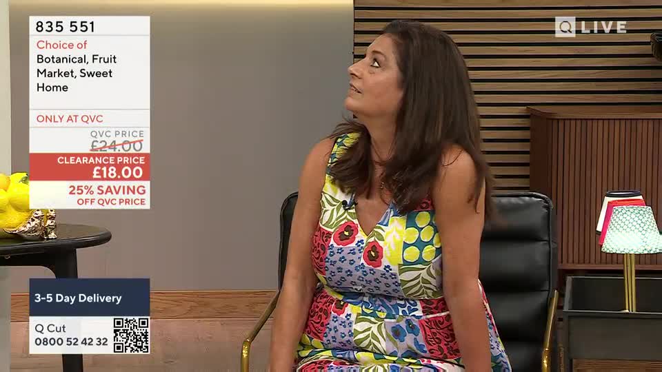 A woman in a colorful dress sits in a chair, looking upwards as if listening. The QVC Extra graphic with the product details is visible on the screen.
