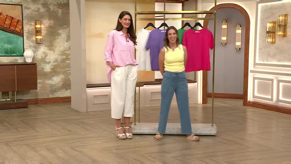 Two women stand in a brightly lit studio, one in pink and white, the other in yellow and blue, both smiling. Behind them, a rack displays colorful t-shirts, likely for a QVC Extra segment in the United Kingdom.
