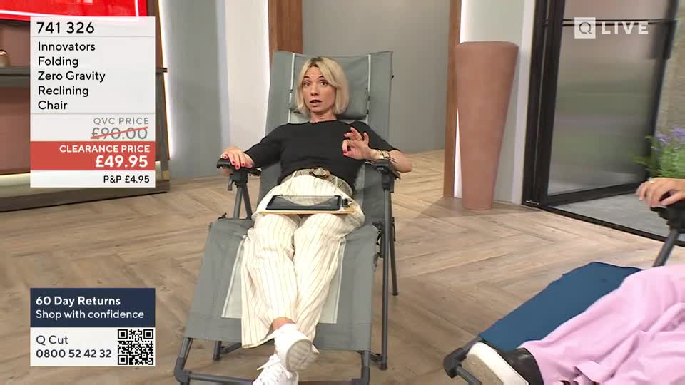 The presenter, seated in a reclining chair, gestures with her right hand while speaking. A tablet rests on a wooden tray across her lap.
