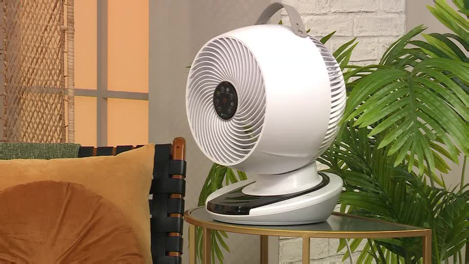 A white fan sits on a small table, its blades designed in a spiral pattern. The QVC Extra set is styled with a tropical plant and a decorative pillow, creating a warm, inviting atmosphere.
