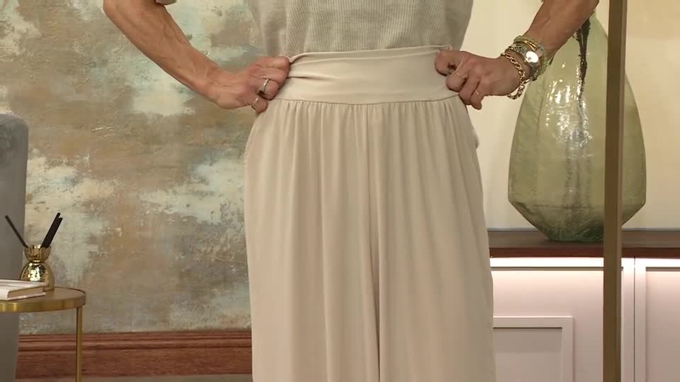 The presenter is holding the waistband of the beige trousers, showing how they sit. The fabric falls in soft, vertical folds from the elasticated waist.
