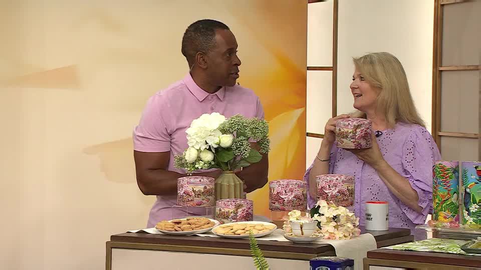 A woman in a purple top holds a decorative tin, smiling as she shows it off. The man beside her, in a pink shirt, is looking at her, and there are more tins and plates of biscuits on the table.
