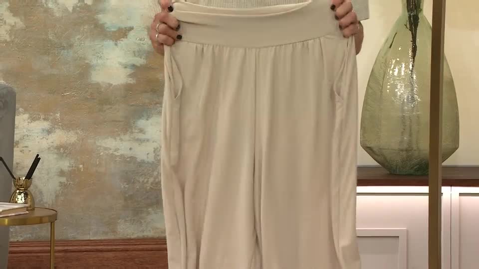 The presenter is holding up a pair of beige trousers, showing the waistband and pockets. The fabric hangs loosely, and the material appears soft.
