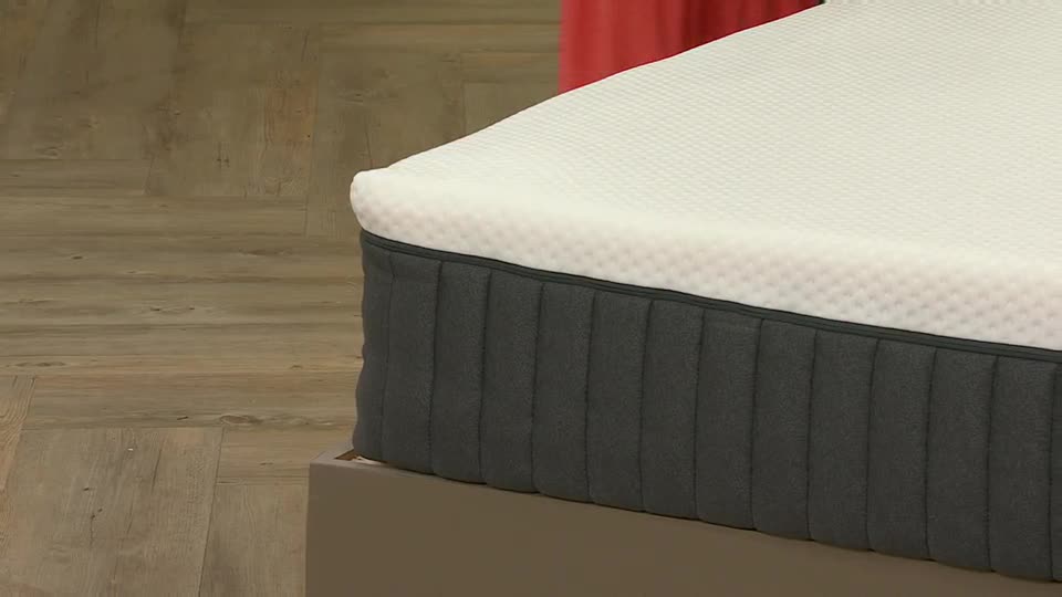 A close-up of a mattress sits on a low platform, the white top layer textured and the gray sides vertically ribbed. The scene looks like it's from QVC Extra in the United Kingdom.
