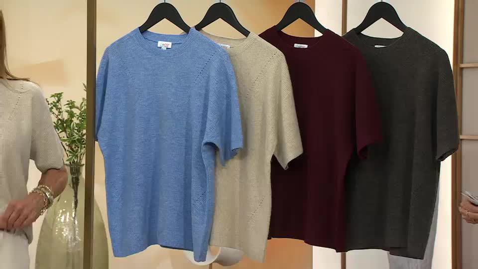 On a QVC Extra set in the United Kingdom, four short-sleeved knit tops hang on a rack, each a different muted color. A model, wearing a similar shirt, stands to the left, while a presenter’s hand gestures towards the garments.
