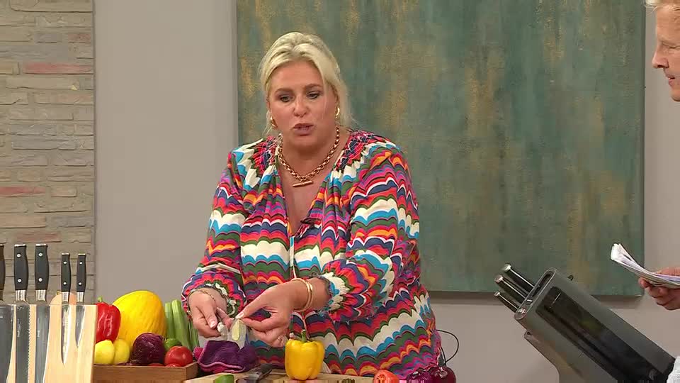 The woman in the colorful top holds a small piece of food, demonstrating something with it. A man off to the side is holding a stack of papers, perhaps a script for the QVC Extra show.
