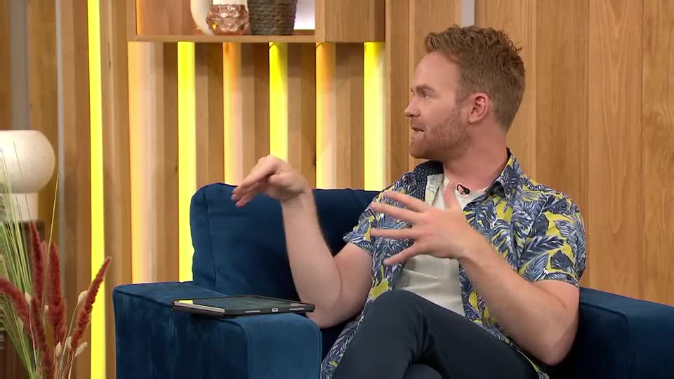 The man, wearing a patterned shirt, gestures with his hands as he speaks. He's sitting on a blue sofa in what looks like a studio, probably QVC Extra in the UK.
