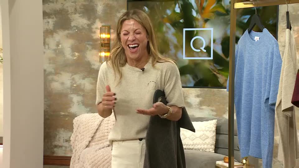 She's laughing, holding a dark sweater and gesturing with her other hand. The set, with its QVC Extra logo, is lit by a wall sconce.
