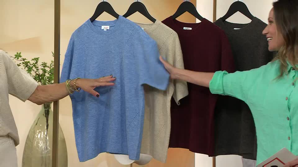 Two women on QVC Extra are gesturing towards a row of short-sleeved sweaters hanging on a rack. One woman is touching the blue one, while the other smiles and points to the next colour.
