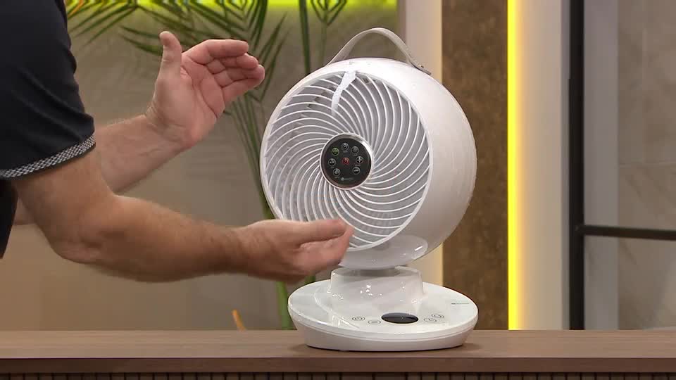 A man gestures towards a white, spherical fan, demonstrating its airflow. The fan's blades are visible, and the presenter seems to be showing how it directs air.
