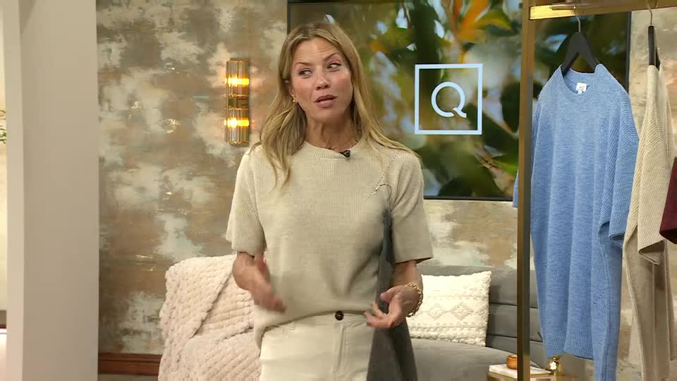 On QVC Extra in the UK, a woman gestures with her hands, presenting a light-colored sweater she's wearing. Behind her, a rack displays other clothing items, including a blue knit dress.
