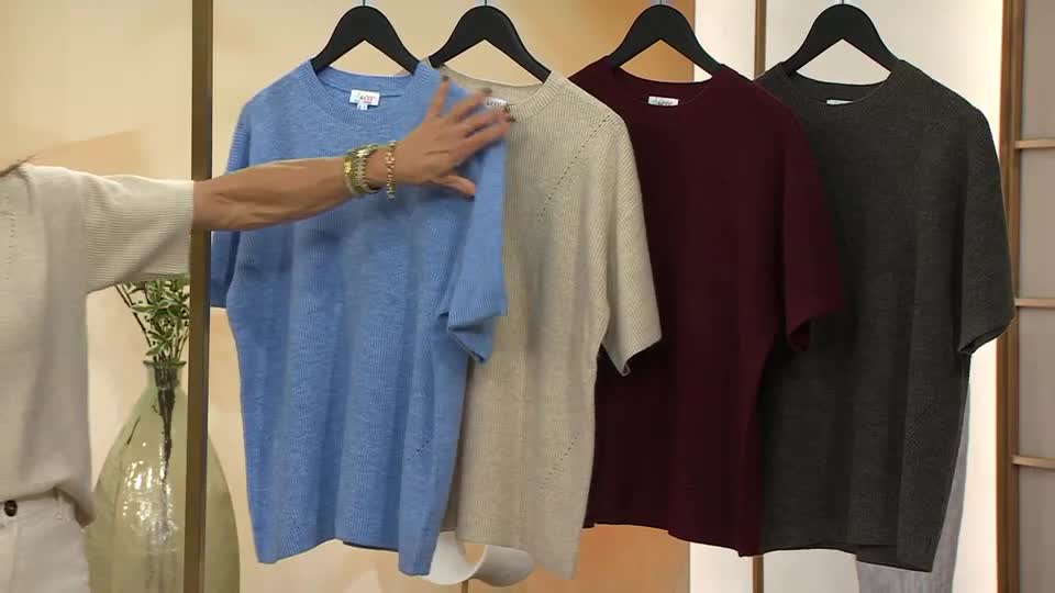 A presenter, on QVC Extra in the United Kingdom, is touching a light blue knit top hanging on a rack. Beside it are tees in beige, burgundy, and gray.
