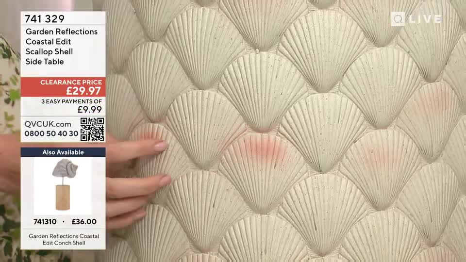 A hand gently strokes the surface of a scallop shell side table, highlighting its textured design. The QVC Extra screen displays the table's price and details.
