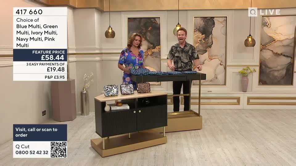 A woman in a floral dress and a man in a patterned shirt stand behind a display table, showcasing patterned bags and fabric. The QVC Extra presenters are in a well-lit studio, likely in the United Kingdom, with a price tag visible on the screen.
