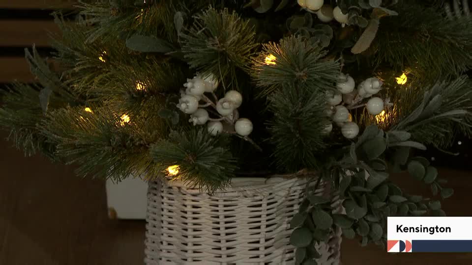 Tiny lights twinkle amongst the faux pine needles and white berries. The arrangement sits in a woven basket, likely a segment on QVC Extra in the United Kingdom.
