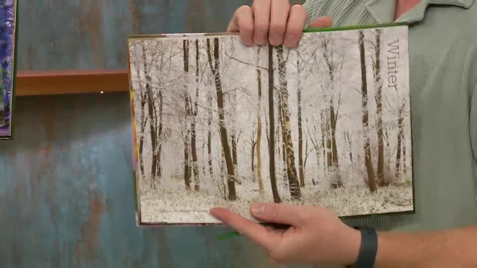 A person is holding open a book, displaying a winter forest scene. The presenter's finger is pointing at the bottom of the page, possibly highlighting a detail.
