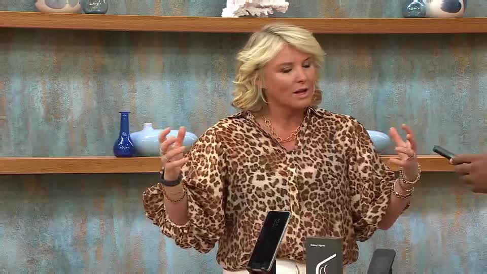 A woman in a leopard-print blouse gestures with both hands, speaking animatedly. A black smartphone is propped up on a stand, and a hand holds another phone off to the side.
