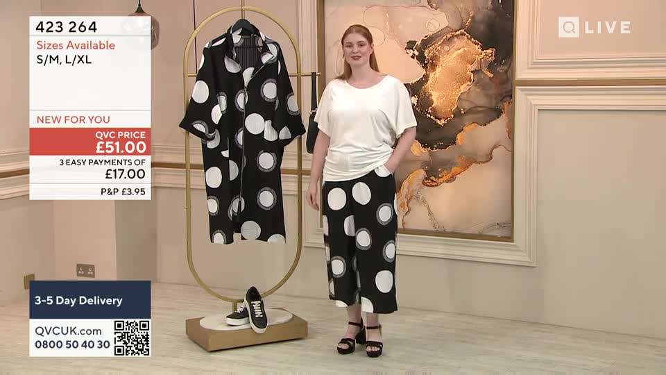 A woman in a white top and polka-dot pants stands center stage on QVC Extra, smiling at the camera.  Behind her, a matching robe hangs on a gold rack, and a pair of sneakers sits on a small platform.
