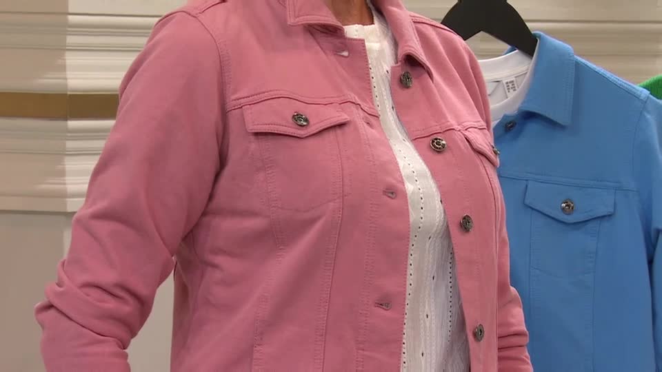 A woman on QVC Extra in the United Kingdom is showing off a rose-colored denim jacket. She's wearing it over a white eyelet top, and a blue jacket hangs behind her on a rack.
