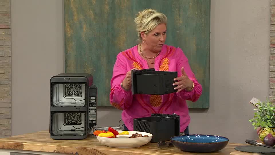 A woman in a pink shirt holds up a black basket, demonstrating it in front of a double-decker air fryer. She's on QVC Extra in the UK, showing off the features of the appliance.
