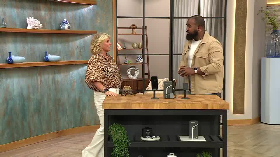 A woman in a leopard-print blouse stands next to a man in a tan jacket, both facing each other. They're positioned behind a dark wood counter, with several black tech items displayed on top.
