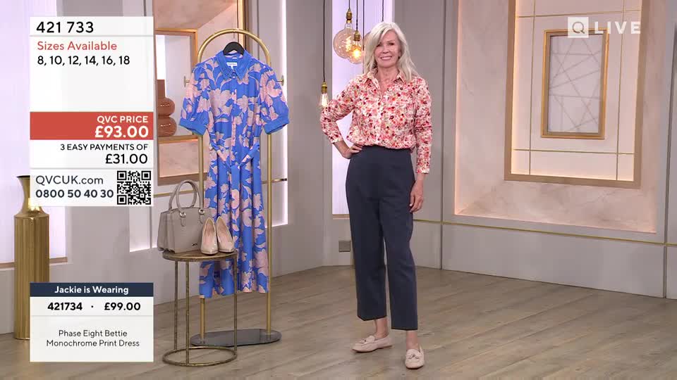 A woman in a floral top and navy trousers stands on a QVC Extra set, smiling with her hand on her hip. Behind her, a blue dress with a floral pattern hangs on a rack, next to a handbag and shoes.
