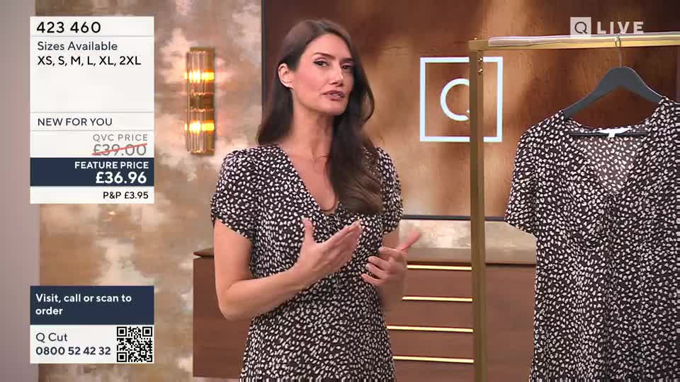 The presenter, wearing the same dress, gestures towards a matching garment hanging on a rack.  She's standing in a studio, likely for QVC Extra in the United Kingdom, with product details displayed beside her.

