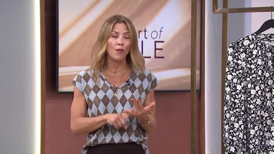The woman in the argyle top gestures with her hands as she speaks, likely on QVC Extra from the United Kingdom. A floral dress hangs on a rack beside her.
