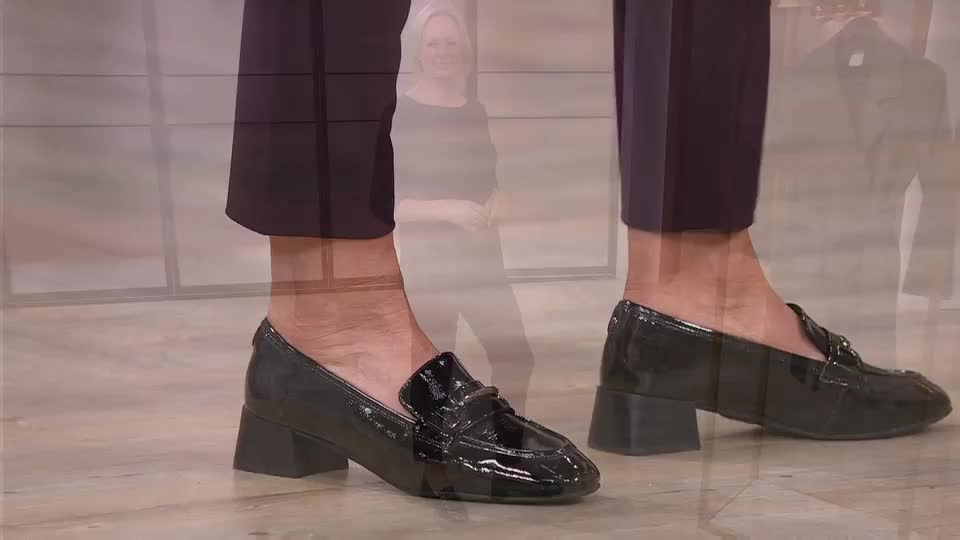 The presenter is showing off shiny black loafers with a small block heel. The camera focuses on the shoes, the trousers, and the presenter's ankles.
