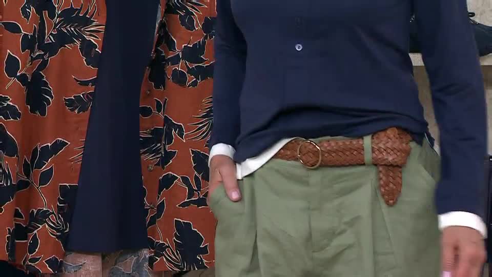 A presenter on QVC Extra in the United Kingdom is showing off a brown braided belt with a gold buckle. The person's hand is in their pocket, and they're wearing a navy blue top over a white shirt.
