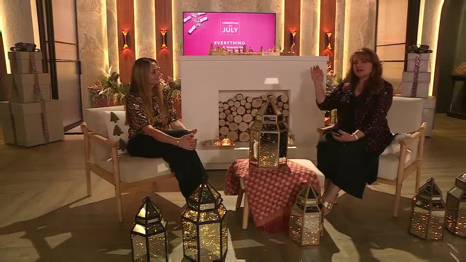 On the QVC Extra set in the UK, two women are seated in armchairs, talking. The woman on the right gestures with her hand, while the other listens intently.
