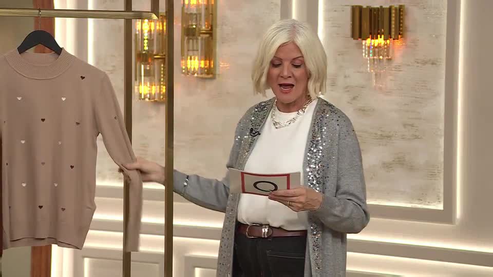 A woman on QVC Extra in the United Kingdom holds up a beige sweater dotted with heart-shaped cutouts. She's reading from a card in her other hand.
