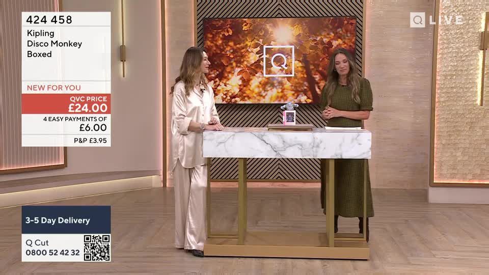 Two women stand at a marble-topped desk, the one on the left in silky pajamas, the other in a green dress. A screen behind them shows a logo, and a graphic displays the price of a "Disco Monkey" item from QVC Extra in the United Kingdom.
