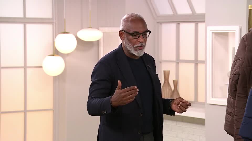 A man in a navy blazer gestures with his hands, speaking to the camera on QVC Extra from the United Kingdom. Behind him, coats hang on a rack, and three globe-shaped lights illuminate the set.
