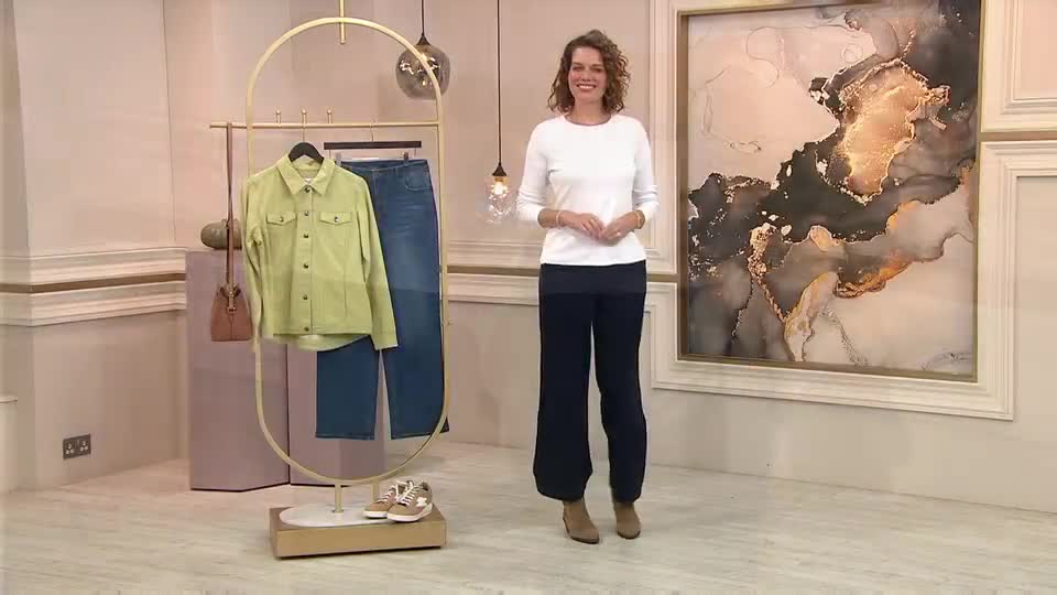 A woman in a white top and dark trousers stands in a well-lit studio, smiling at the camera. Behind her, a clothing rack holds a chartreuse jacket and jeans, and a large abstract painting hangs on the wall.
