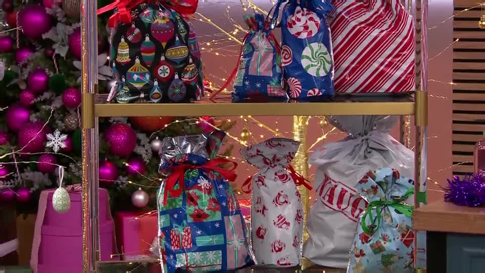 On a shelf, colorful gift bags are arranged, ready for the holiday season. The bags, with patterns like ornaments and candy canes, are tied with ribbons and sit in front of a decorated Christmas tree.
