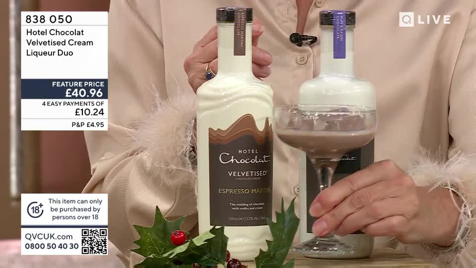 A presenter holds two bottles of Hotel Chocolat liqueur, one in each hand, while a cocktail glass is filled with a creamy, brown liquid. The QVC Extra set features a festive arrangement of holly and berries.
