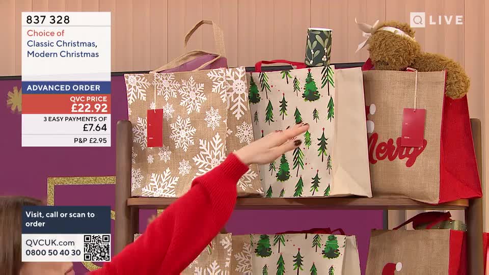 A woman in a red sweater gestures towards a row of Christmas-themed tote bags on a shelf. The bags, with festive designs, are part of a QVC Extra presentation in the United Kingdom.
