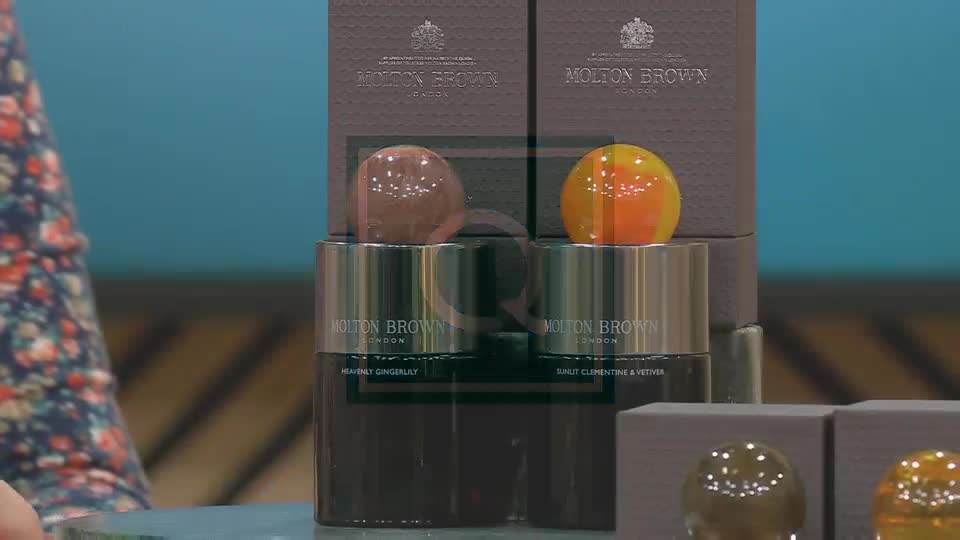 Two Molton Brown bottles, one with a brown orb and the other with a yellow one, are displayed on QVC Extra. The presenter's floral sleeve is visible in the corner, and the background is a solid turquoise.
