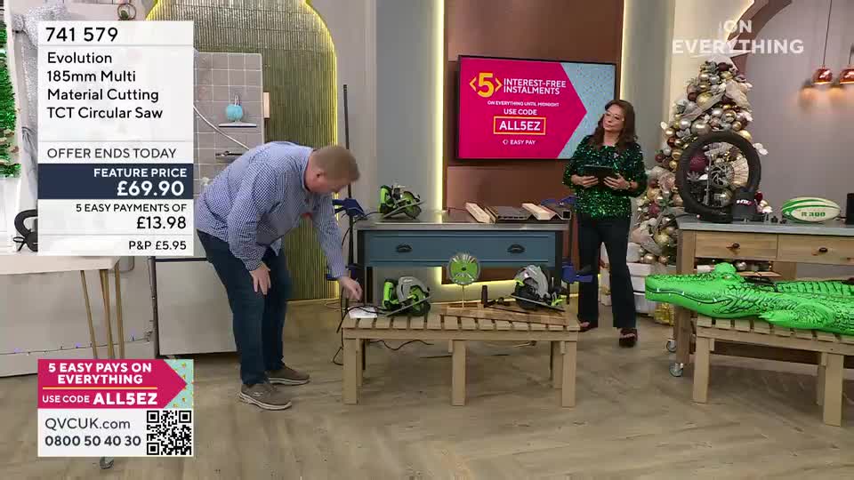 A man in a blue shirt is bent over, fiddling with a power cord on a table. Across the studio, a woman in a green sequined top holds a tablet, looking towards the camera.
