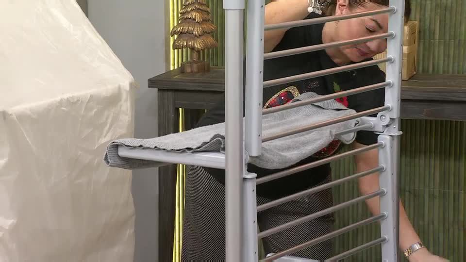 A woman in a black t-shirt carefully places a grey sweater onto a drying rack. She's demonstrating the product on QVC Extra from the United Kingdom, and the studio set looks cozy.
