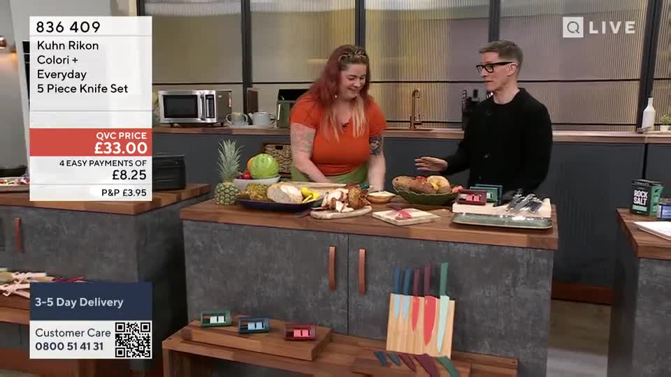 A woman in an orange top and a man in a black sweater stand behind a kitchen counter, gesturing towards the food and knives laid out. The QVC Extra set features colorful knives and a display of food, likely for a product demonstration.
