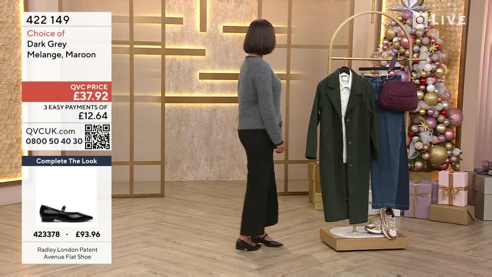 A woman in a grey sweater and black pants stands near a rack of clothing on the QVC Extra set. Behind her, a Christmas tree glitters, and a screen displays product information.
